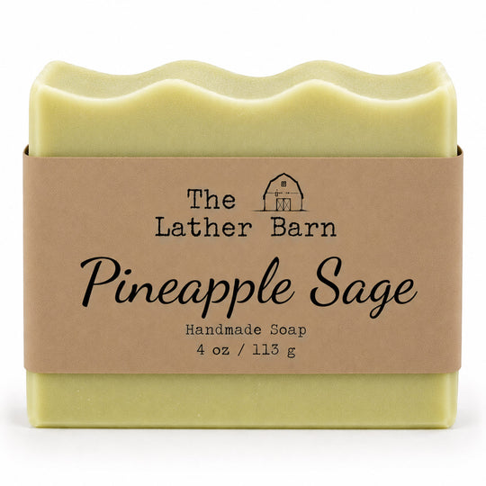 A 4 oz bar of handmade soap by The Lather Barn, named Pineapple Sage. The soap is pale yellow-green and wrapped in a brown kraft paper band with black text and a small barn logo, set against a plain white background.