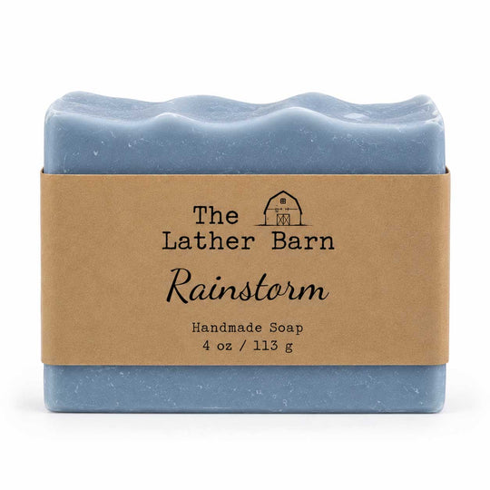 A rectangular bar of handmade soap in a dusty light blue color, wrapped in a brown kraft paper band with the brand name 'The Lather Barn' and product name 'Rainstorm' printed on it, set against a plain white background.