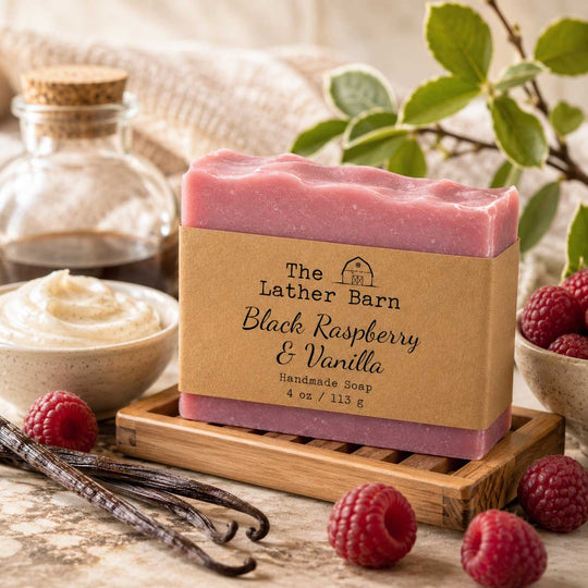 A bar of handmade soap labeled 'The Lather Barn Black Raspberry & Vanilla Handmade Soap' sits on a wooden soap dish, surrounded by fresh raspberries, vanilla beans, a bowl of whipped cream, and a glass jar in a styled setting.