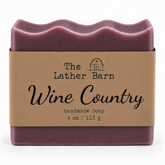 A single bar of handmade soap wrapped in a kraft paper band. The soap has a deep wine-red color with a wavy top edge, and the label reads 'The Lather Barn Wine Country Handmade Soap 4 oz / 113 g'.