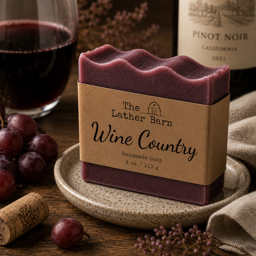 A bar of handmade soap labeled 'The Lather Barn Wine Country' sits on a small ceramic dish on a wooden table, styled with grapes, a wine bottle, a glass of red wine, and a cork.