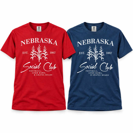 Two short-sleeve graphic T-shirts shown side by side on a white background: one red and one navy blue. Each shirt has a white printed design reading "NEBRASKA Social Club" with corn graphics and smaller text about football, corn bowls, and gravel roads.