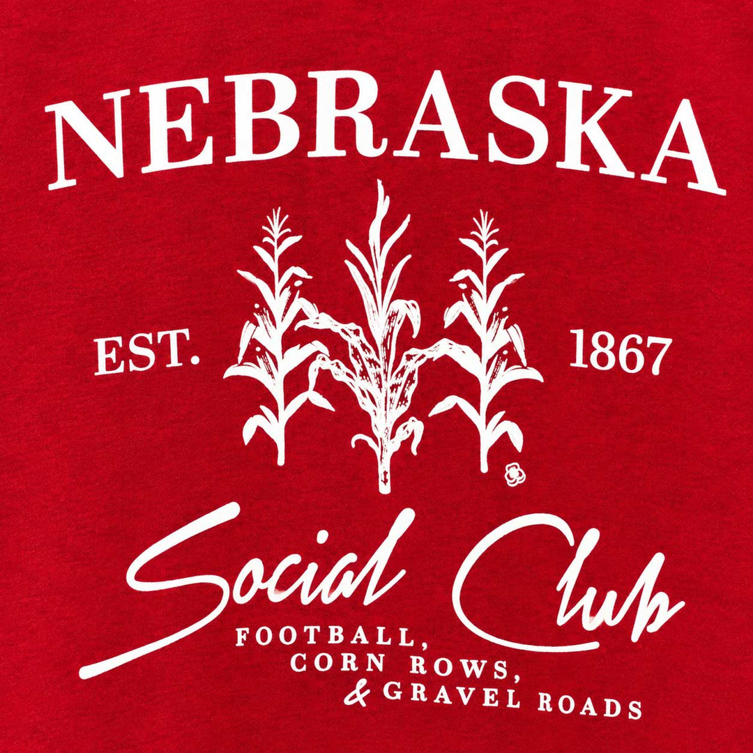 A close-up of a red graphic print featuring the word 'NEBRASKA' in large white letters above three corn silhouettes, with 'EST. 1867' on either side and 'Social Club' in white script below, along with smaller text reading 'FOOTBALL, CORN ROWS, & GRAVEL ROADS.