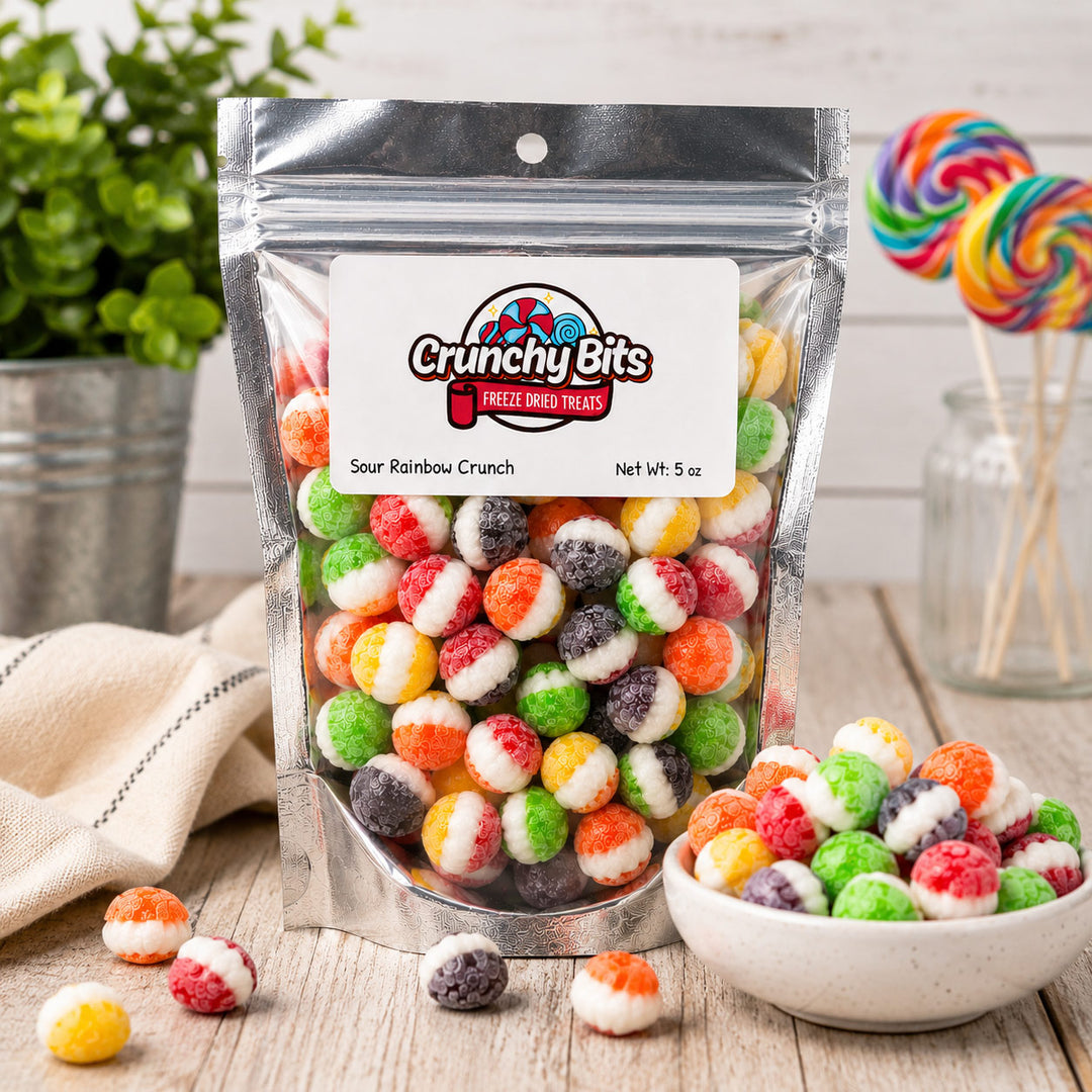 A stand-up pouch of Crunchy Bits Sour Rainbow Crunch frozen dried treats on a rustic wooden table, with a small bowl of the candy pieces and colorful lollipops in the background.