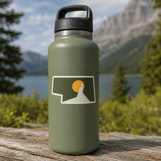 An olive green insulated water bottle with a black lid sits on a weathered wooden log outdoors, with a blurred mountain and lake landscape in the background. The bottle features a white graphic of a mountain peak with a yellow sun inside a speech-bubble outline.