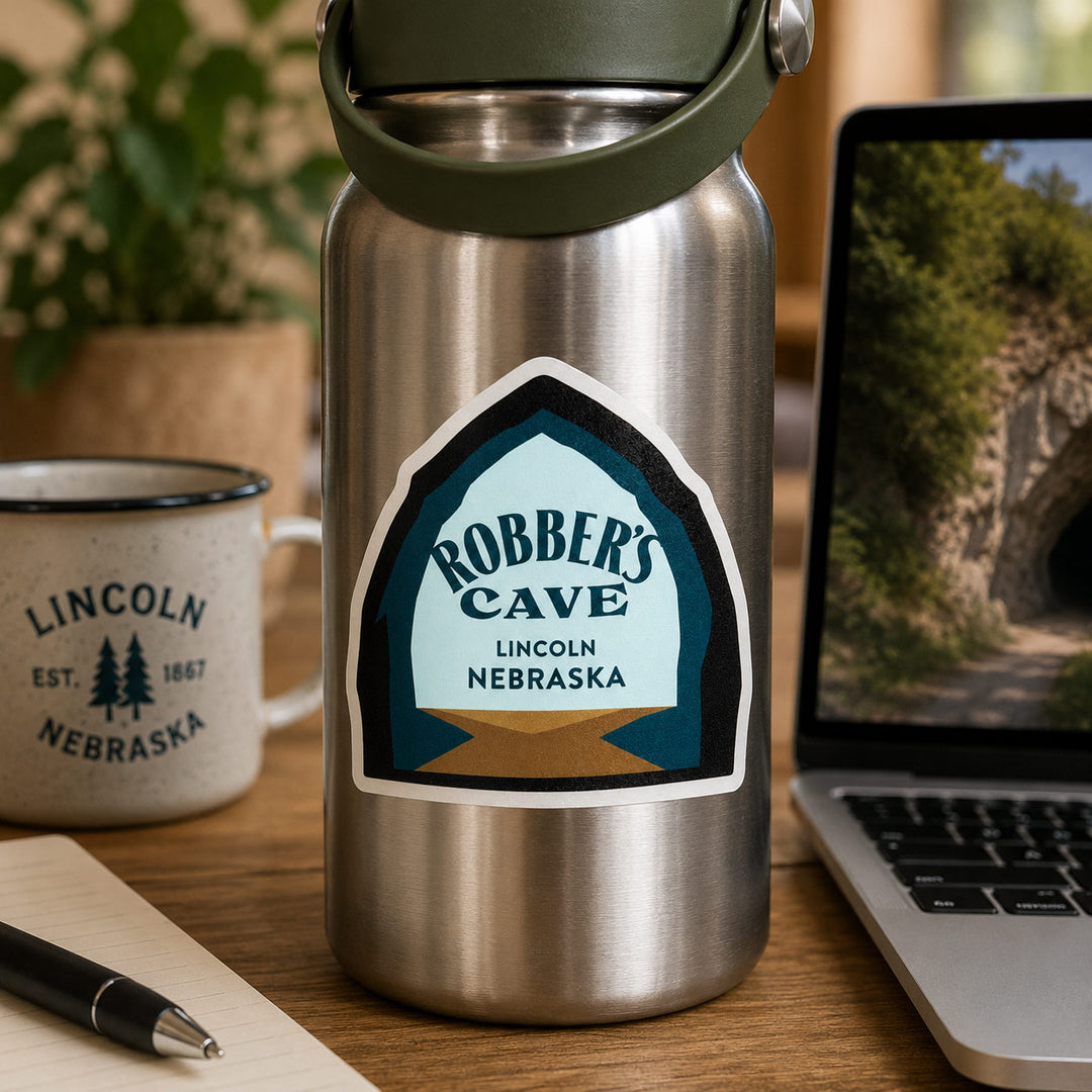 A stainless steel insulated water bottle with a dark green lid and carry loop sits on a wooden desk. The bottle has a large decorative sticker reading 'Robber's Cave Lincoln Nebraska.
