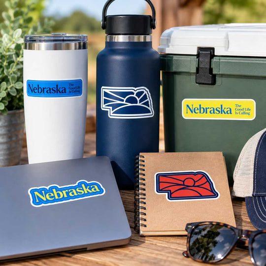 A set of Nebraska-themed decals displayed on various everyday items including a laptop, tumbler, water bottle, cooler, notebook, and cap, arranged on a wooden table outdoors.