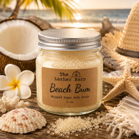 A glass jar of whipped sugar body scrub labeled 'The Lather Barn Beach Bum' sits on a wooden surface surrounded by beach-themed props including a coconut shell, flower, starfish, seashells, and a straw hat, with a blurred beach background.