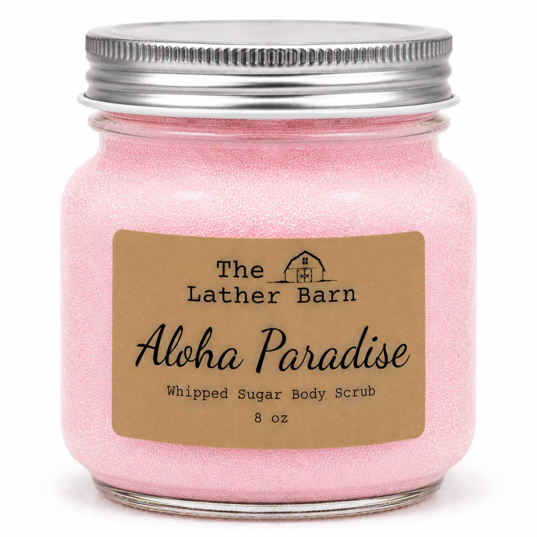 A pink whipped sugar body scrub in a clear glass jar with a silver metal lid and a brown paper label reading 'The Lather Barn Aloha Paradise Whipped Sugar Body Scrub 8 oz'.