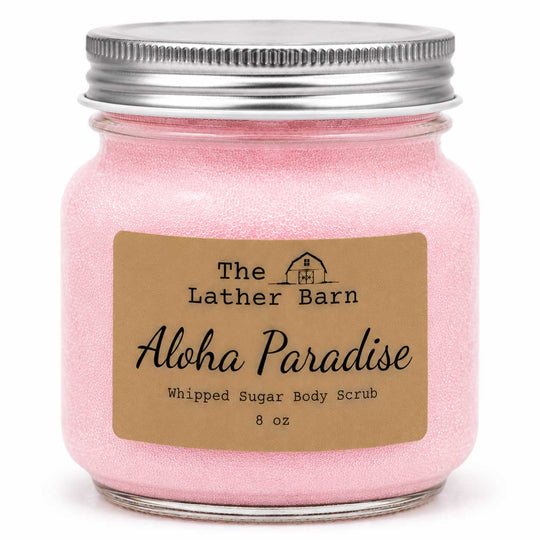 A pink whipped sugar body scrub in a clear glass jar with a silver metal lid and a brown paper label reading 'The Lather Barn Aloha Paradise Whipped Sugar Body Scrub 8 oz'.