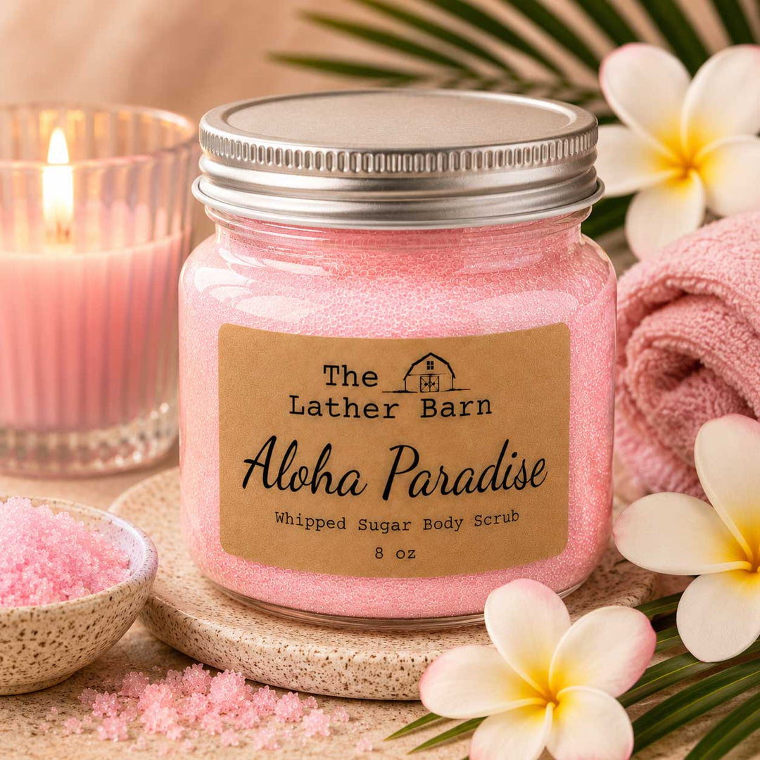 A jar of pink whipped sugar body scrub by The Lather Barn, labeled 'Aloha Paradise,' styled with a pink candle, plumeria flowers, a towel, and scattered scrub particles on a warm neutral background.