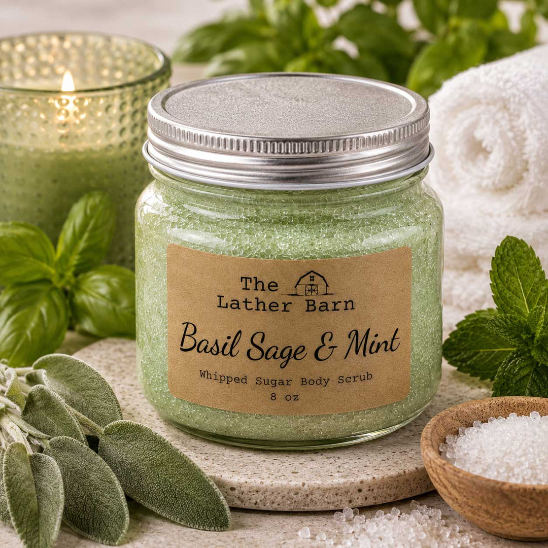 A jar of whipped sugar body scrub by The Lather Barn, labeled 'Basil Sage & Mint,' filled with pale green scrub and topped with a silver lid. The jar is styled on a stone coaster with mint leaves, coarse salt, a lit candle, and a rolled towel in the background.