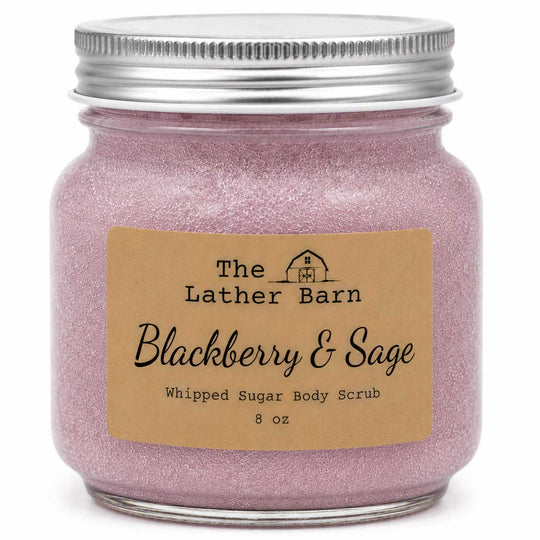 An 8 oz glass jar candle or body scrub by The Lather Barn, featuring a silver metal lid and a kraft paper label that reads 'Blackberry & Sage Whipped Sugar Body Scrub'. The contents are a sparkling light pink-lilac color.