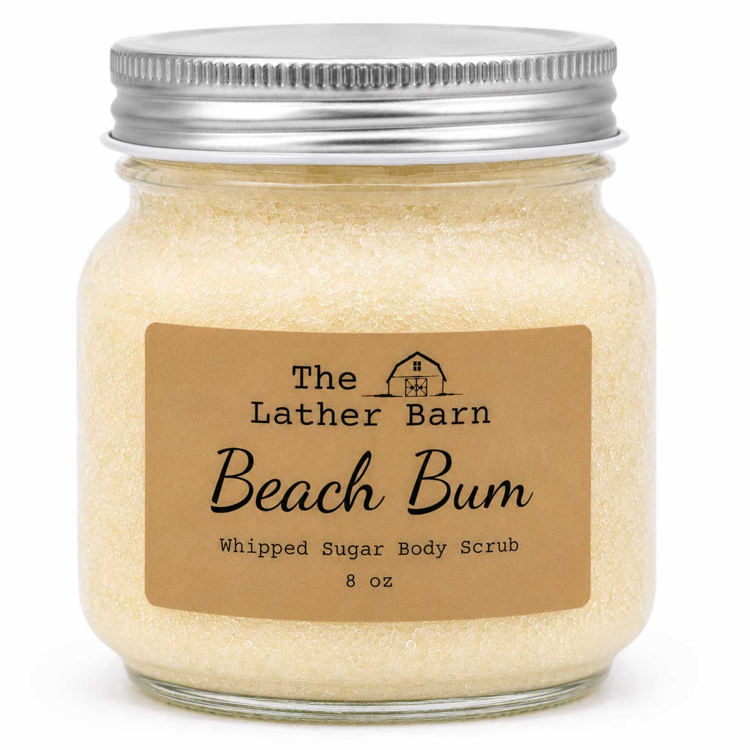 An 8 oz glass jar of 'Beach Bum' whipped sugar body scrub by The Lather Barn, filled with pale yellow-beige scrub and sealed with a silver metal lid, set against a plain white background.