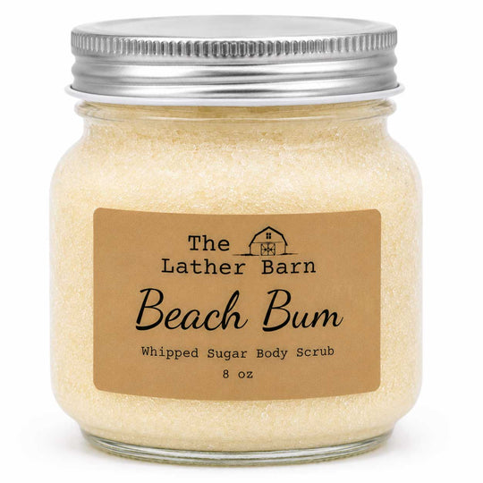An 8 oz glass jar of 'Beach Bum' whipped sugar body scrub by The Lather Barn, filled with pale yellow-beige scrub and sealed with a silver metal lid, set against a plain white background.