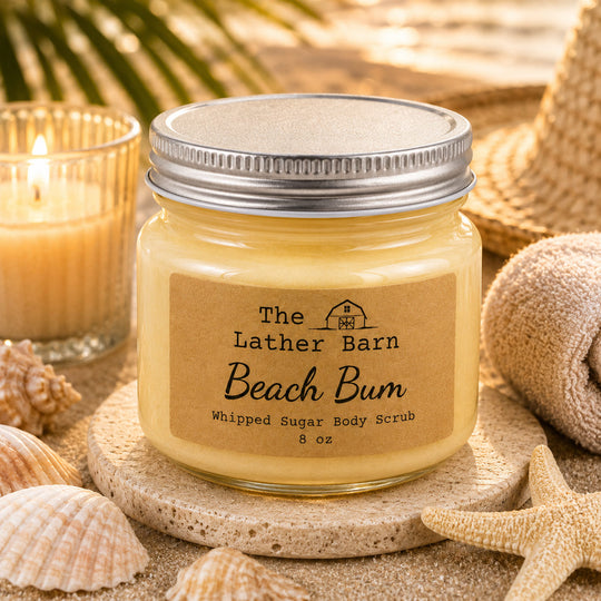 A jar of whipped sugar body scrub labeled 'Beach Bum' by The Lather Barn, styled on a stone coaster with seashells, sand, a candle, and a straw hat in a beach-themed setting.