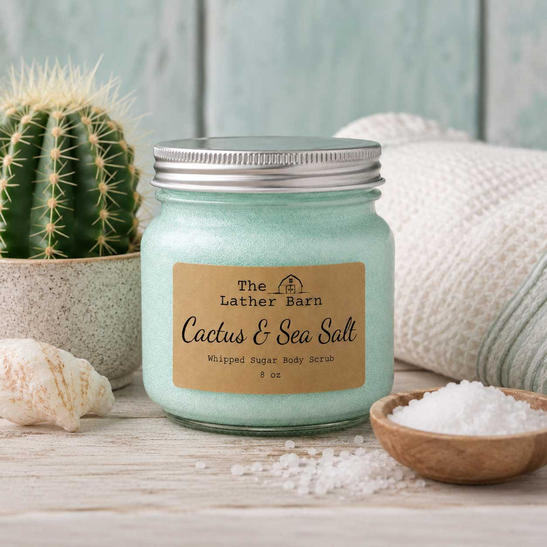 A sugar scrub labeled 'The Lather Barn Cactus & Sea Salt' sits on a rustic wooden surface with a cactus, shell, bowl of white salt, and soft textiles in the background.
