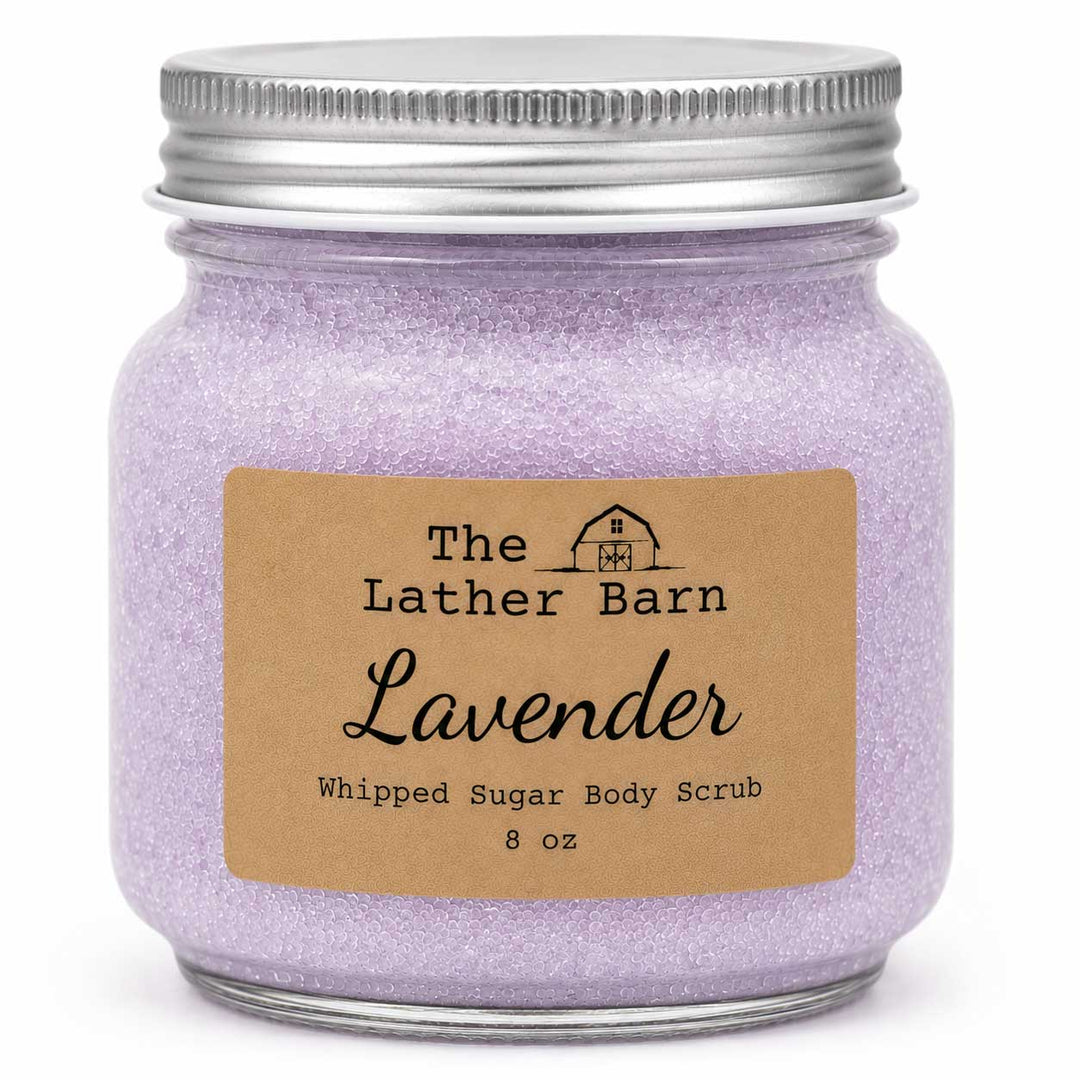 A jar of lavender whipped sugar body scrub by The Lather Barn, featuring a silver metal screw-top jar filled with light purple glittery scrub and a brown kraft paper label.