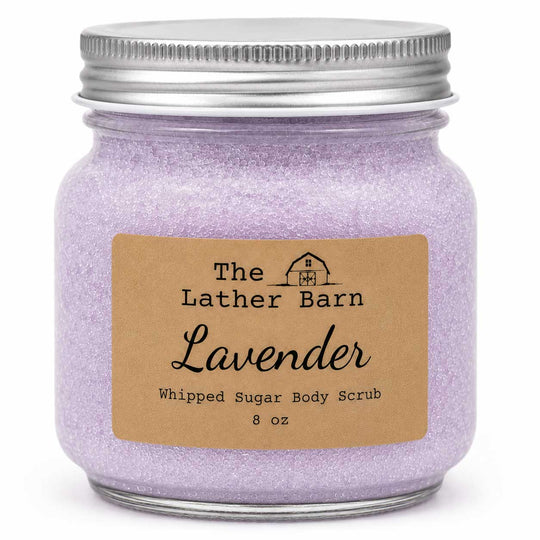 A jar of lavender whipped sugar body scrub by The Lather Barn, featuring a silver metal screw-top jar filled with light purple glittery scrub and a brown kraft paper label.
