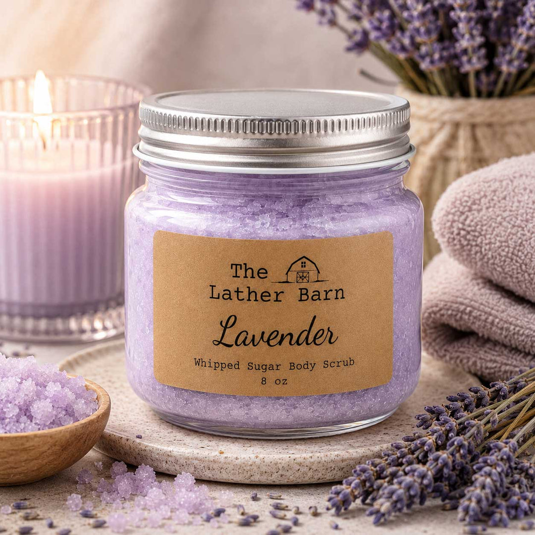 A glass jar of lavender whipped sugar body scrub by The Lather Barn, styled with lavender sprigs, a lit candle, and a folded towel.