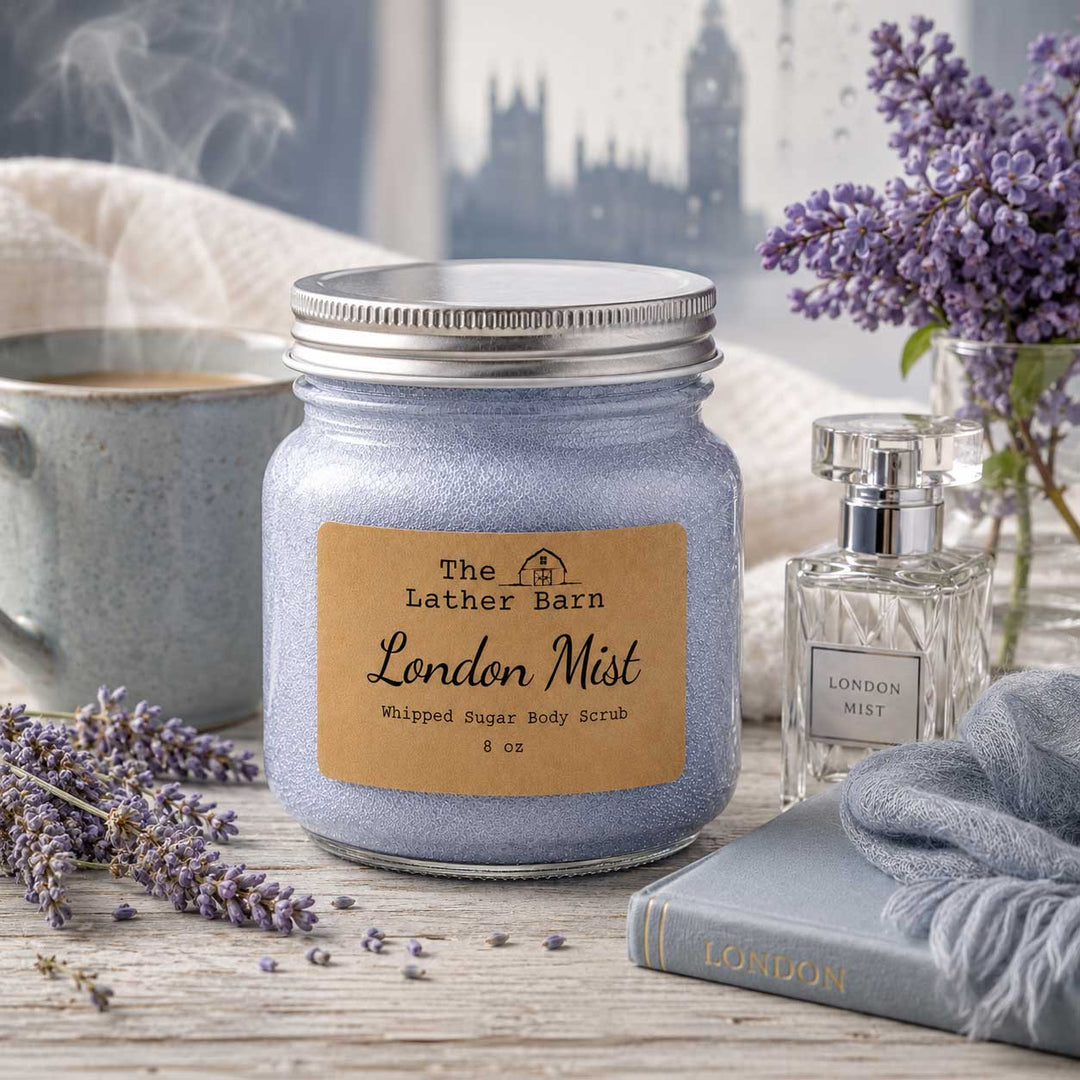 A jar candle labeled 'The Lather Barn London Mist' sits on a rustic wooden surface with lavender flowers, a gray mug, a perfume bottle, and a gray scarf in a styled setting.