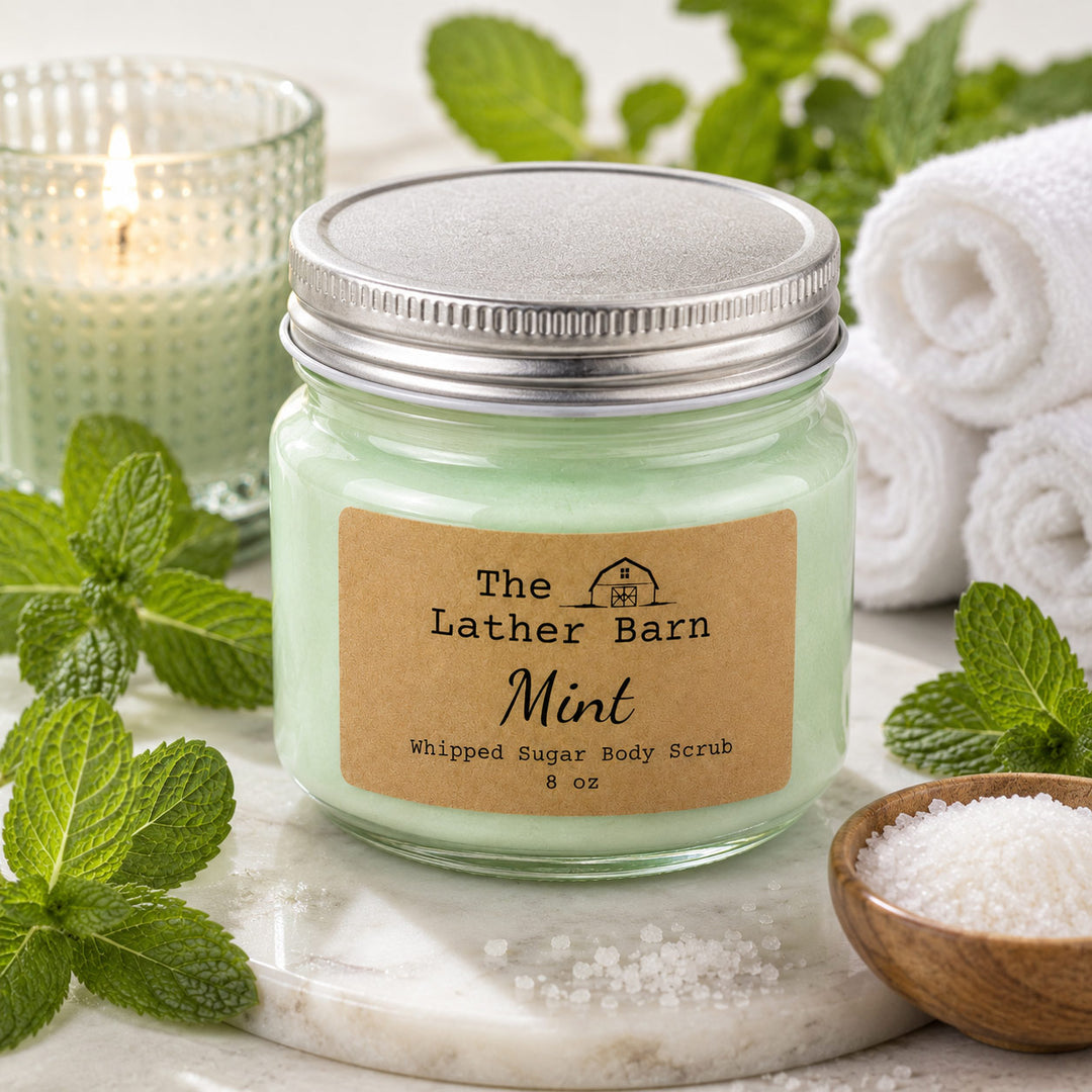 A jar of whipped sugar body scrub by The Lather Barn, labeled 'Mint', styled on a marble slab with mint leaves, a lit candle, rolled towels, and a small bowl of sea salt.