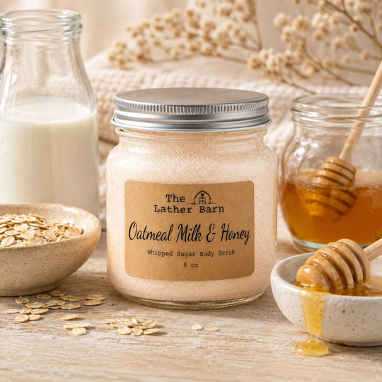 A jar of oatmeal milk and honey whipped sugar body scrub by The Lather Barn, styled on a rustic wooden surface with oats, a glass bottle of milk, honey, and a honey dipper.
