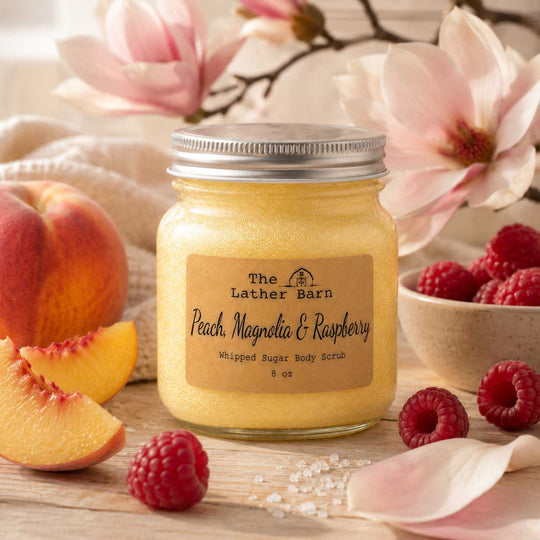 A glass jar of whipped sugar body scrub by The Lather Barn, labeled 'Peach, Magnolia & Raspberry,' styled on a wooden surface with peach slices, whole raspberries, a bowl of raspberries, and soft pink flowers in the background.