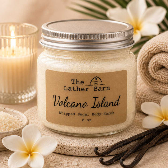 A jar of whipped sugar body scrub by The Lather Barn, labeled 'Volcano Island,' styled with white flowers, vanilla beans, a lit candle, and a rolled towel on a spa-like surface.
