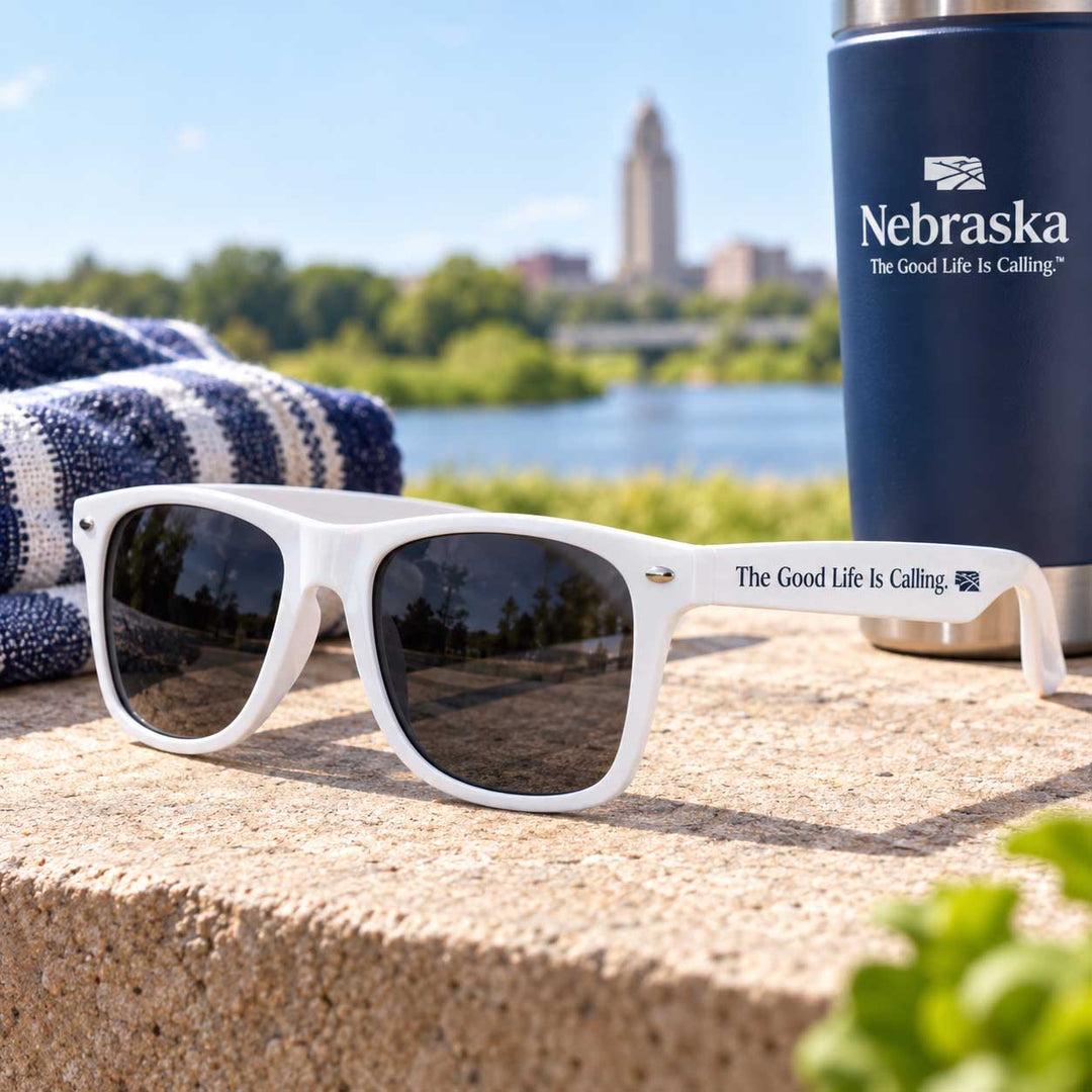 White sunglasses with dark lenses resting on a stone surface outdoors, with a blue Nebraska-branded tumbler and a striped towel in the background near a river and buildings.