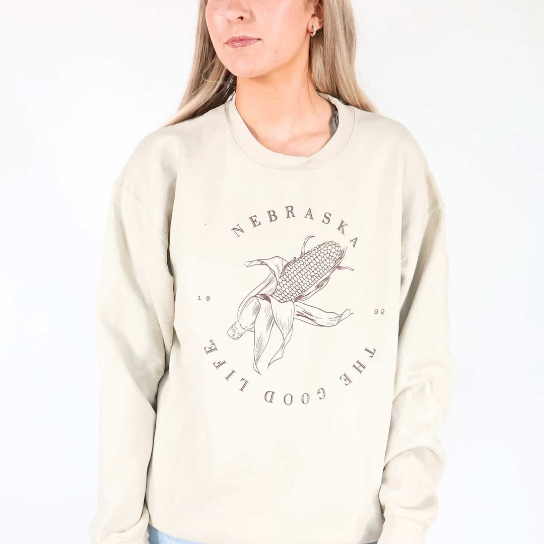 A woman wearing a cream-colored crewneck sweatshirt with a brown graphic of a corn cob and the text 'NEBRASKA THE GOOD LIFE' printed on the front, set against a plain white background.
