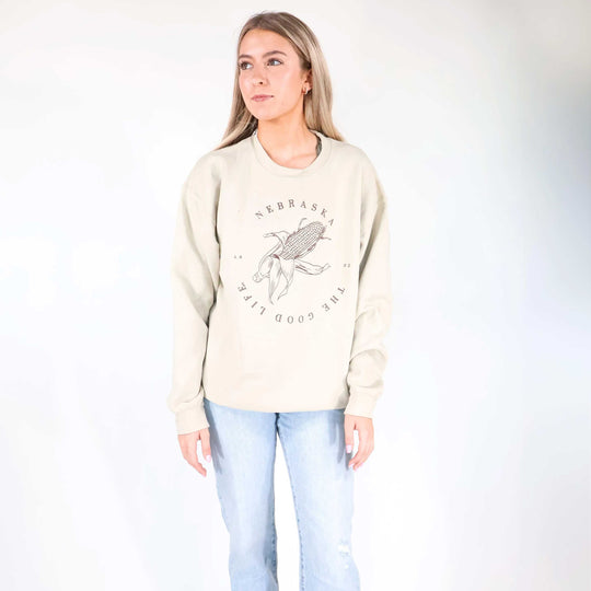 A woman modeling a cream-colored crewneck sweatshirt with a black Nebraska corn graphic and text, paired with light blue jeans, against a plain white studio background.