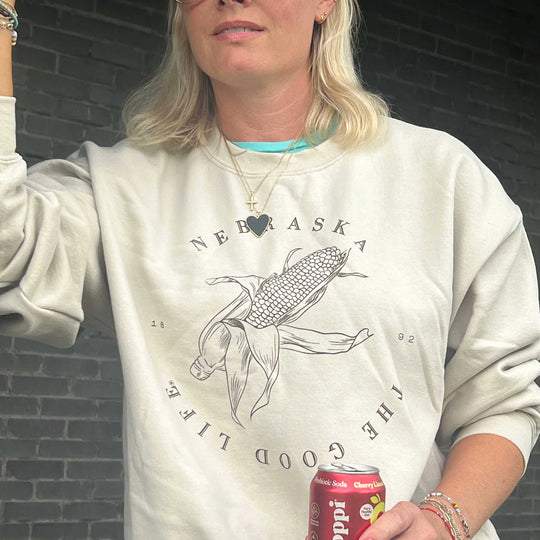 A woman wearing a cream-colored sweatshirt with a black line art graphic of a corn cob and the words 'NEBRASKA' and 'THE GOOD LIFE'. She is holding a red Pepsi can against a dark gray brick wall background.