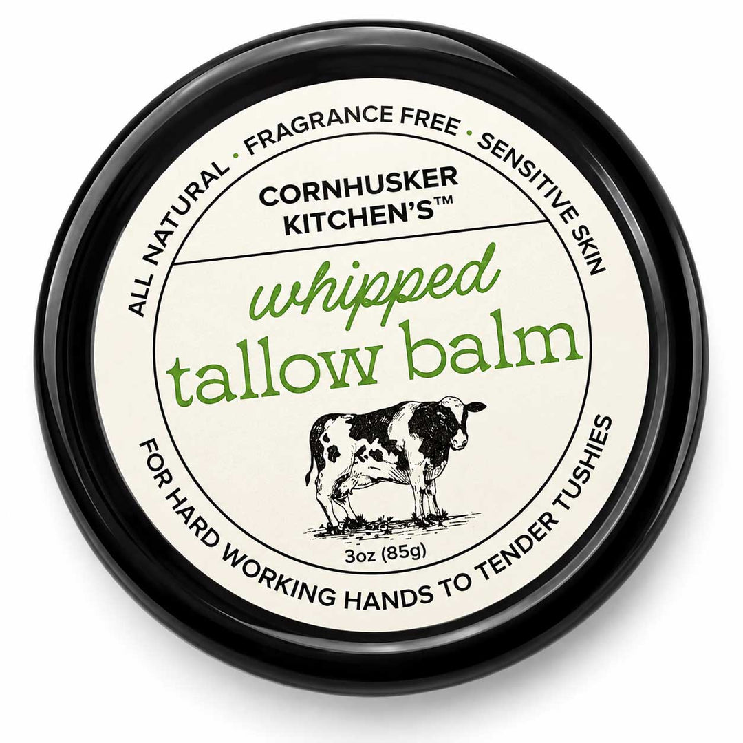 A round black tin of Cornhusker Kitchen's Whipped Tallow Balm, featuring a cream-colored label with green and black text and an illustration of a cow.