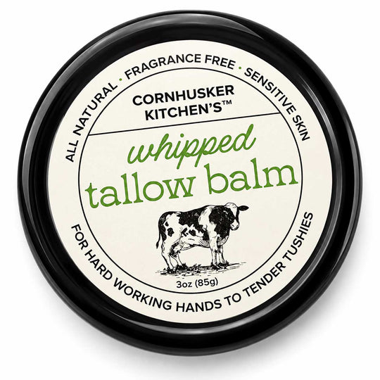 A round black tin of Cornhusker Kitchen's Whipped Tallow Balm, featuring a cream-colored label with green and black text and an illustration of a cow.