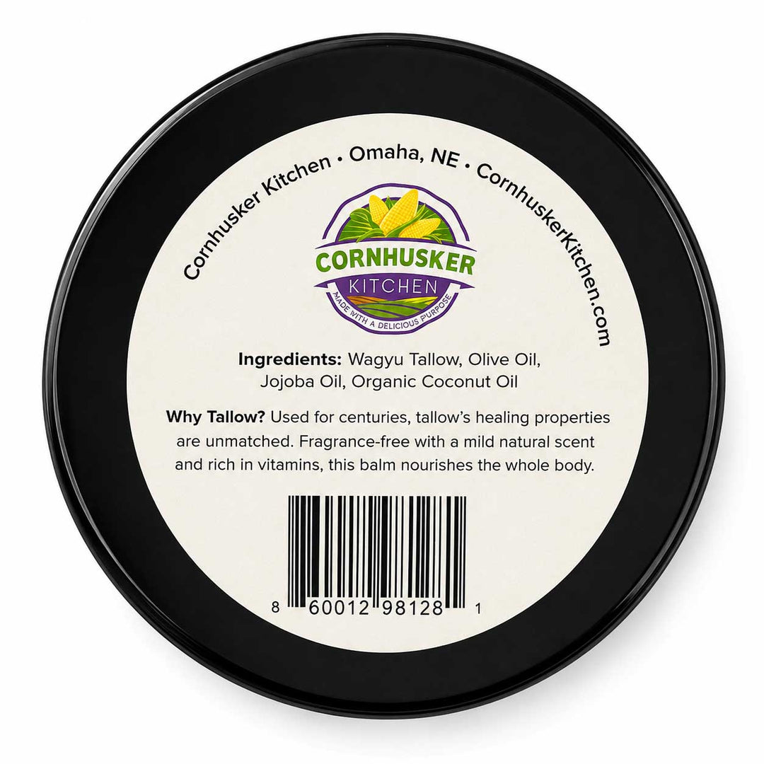 Top-down view of a round black plastic container lid with a white label for Cornhusker Kitchen Tallow balm, showing the brand logo, ingredients, and barcode.