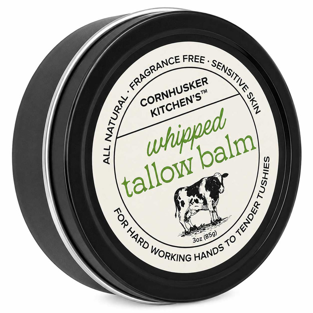 A round black tin of Cornhusker Kitchen's Whipped Tallow Balm, featuring a cream-colored label with green and black text and an illustration of a cow.