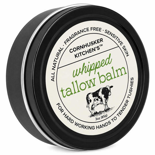 A round black tin of Cornhusker Kitchen's Whipped Tallow Balm, featuring a cream-colored label with green and black text and an illustration of a cow.