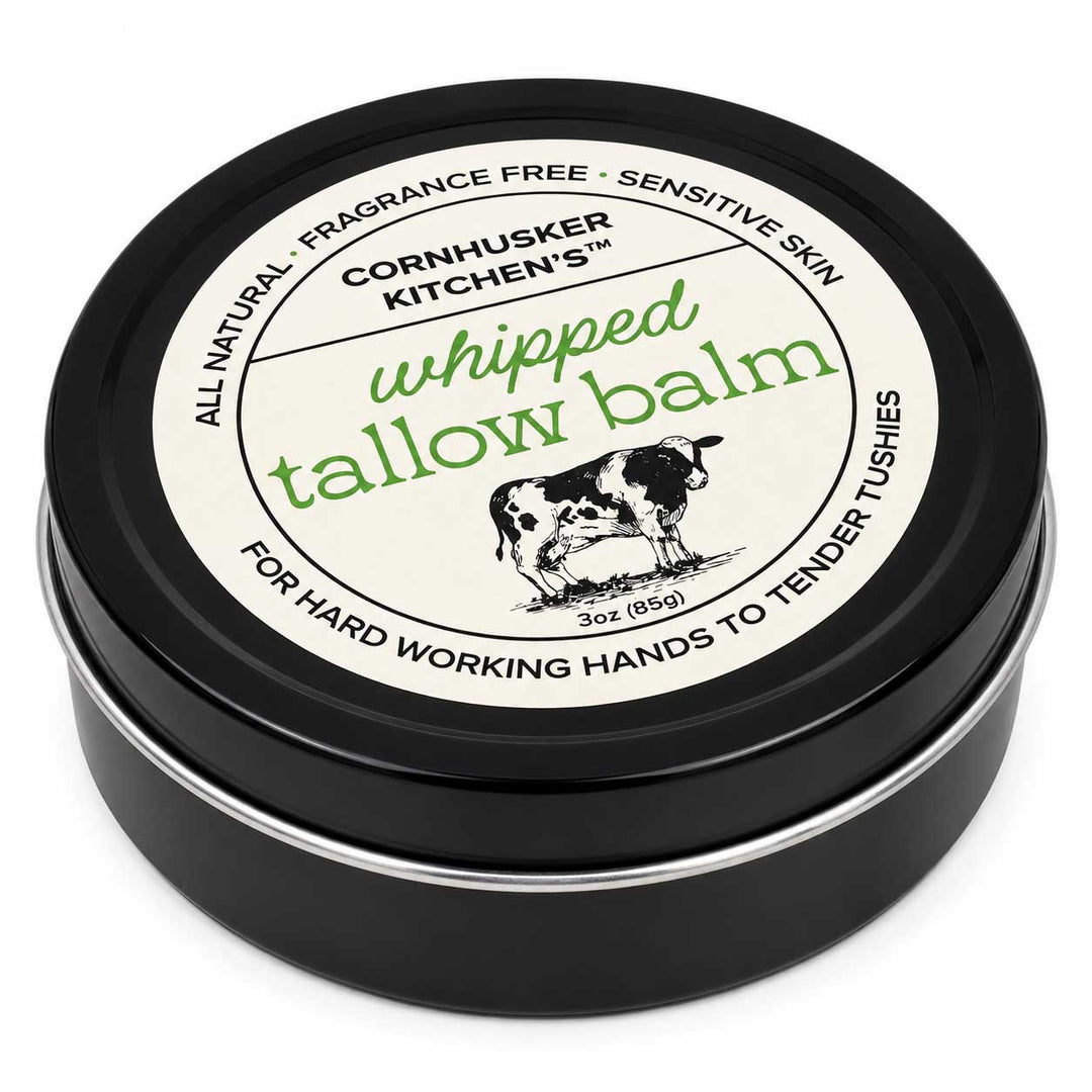 A round black tin of Cornhusker Kitchen's Whipped Tallow Balm, featuring a cream-colored label with green and black text and an illustration of a cow.