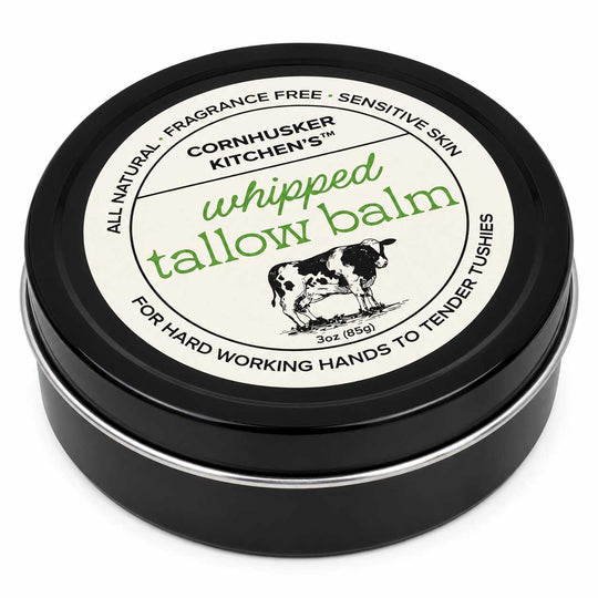 A round black tin of Cornhusker Kitchen's Whipped Tallow Balm, featuring a cream-colored label with green and black text and an illustration of a cow.