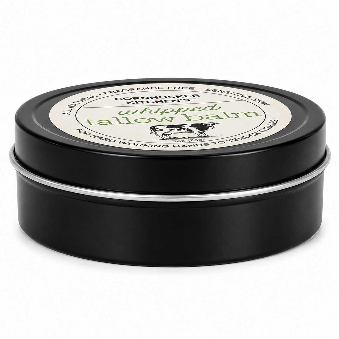 A round black metal tin of whipped tallow balm with a white label on top, photographed on a clean white background.
