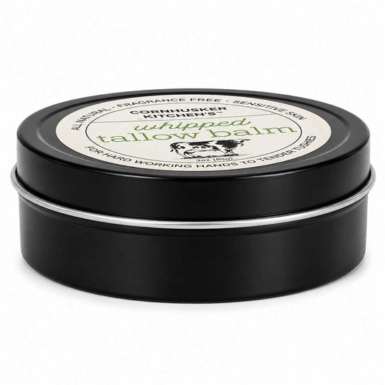 A round black metal tin of whipped tallow balm with a white label on top, photographed on a clean white background.