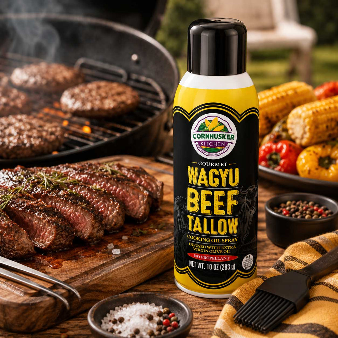 A bottle of Cornhusker Kitchen Wagyu Beef Tallow Cooking Oil Spray stands on a wooden table in a barbecue setting, with sliced beef, grill marks, corn, peppers, salt, peppercorns, and a basting brush around it.