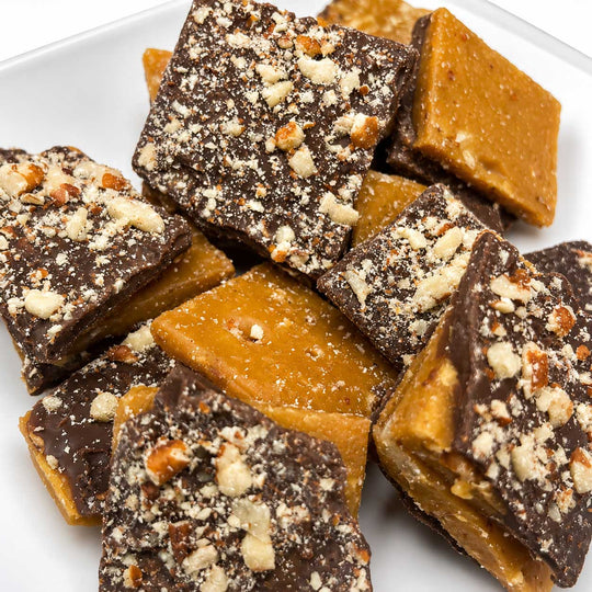 A pile of square caramel and chocolate bark pieces on a white plate, topped with chopped nuts and coarse sugar crystals.