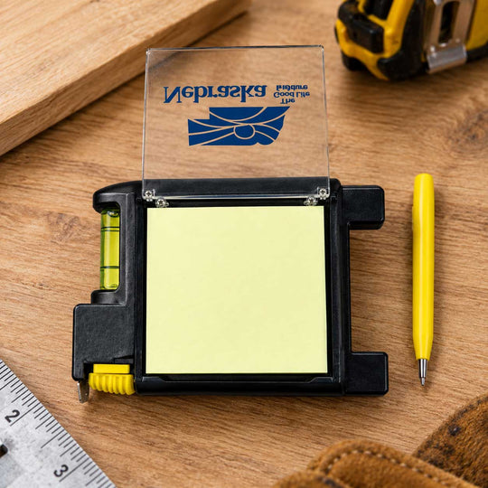 A Nebraska temporary wood tape measure with a clear cover and yellow sticky note pad, shown on a wooden workbench with a yellow pen and measuring tools nearby.