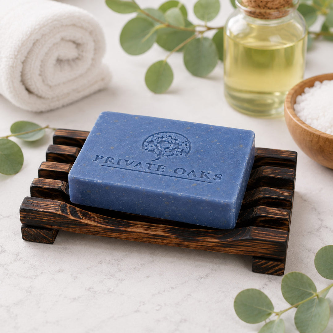 A rectangular blue soap bar embossed with the brand name 'PRIVATE OAKS' sits on a dark wooden soap dish. The scene is styled with a rolled white towel, a small bottle of oil, a bowl of salt, and green eucalyptus leaves on a light marble surface.