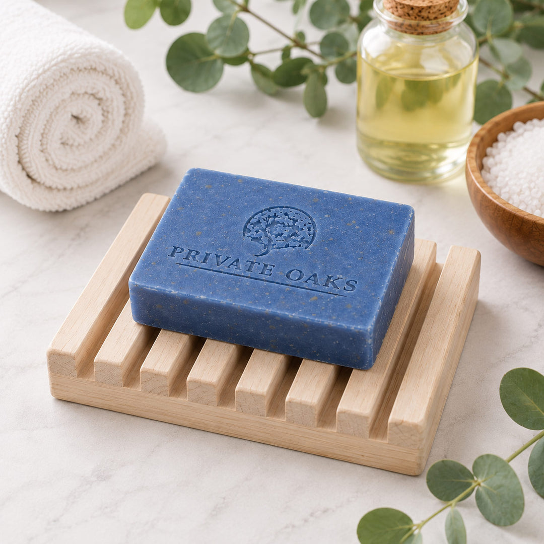 A rectangular blue soap bar embossed with the brand name 'PRIVATE OAKS' sits on a light natural wood soap dish. The scene is styled with a rolled white towel, a small glass bottle of oil, a bowl of white salt, and green eucalyptus leaves on a light marble surface.