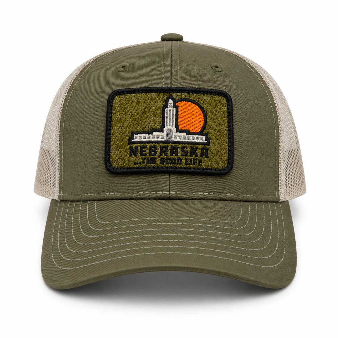 An olive green and beige trucker hat featuring a rectangular embroidered patch on the front with the Nebraska statehouse and a sunset, reading 'NEBRASKA .