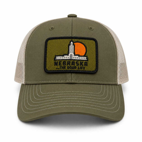 An olive green and beige trucker hat featuring a rectangular embroidered patch on the front with the Nebraska statehouse and a sunset, reading 'NEBRASKA .
