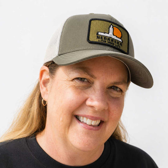 A smiling woman wearing a gray trucker hat with a Nebraska flag patch on the front, photographed against a plain light background.